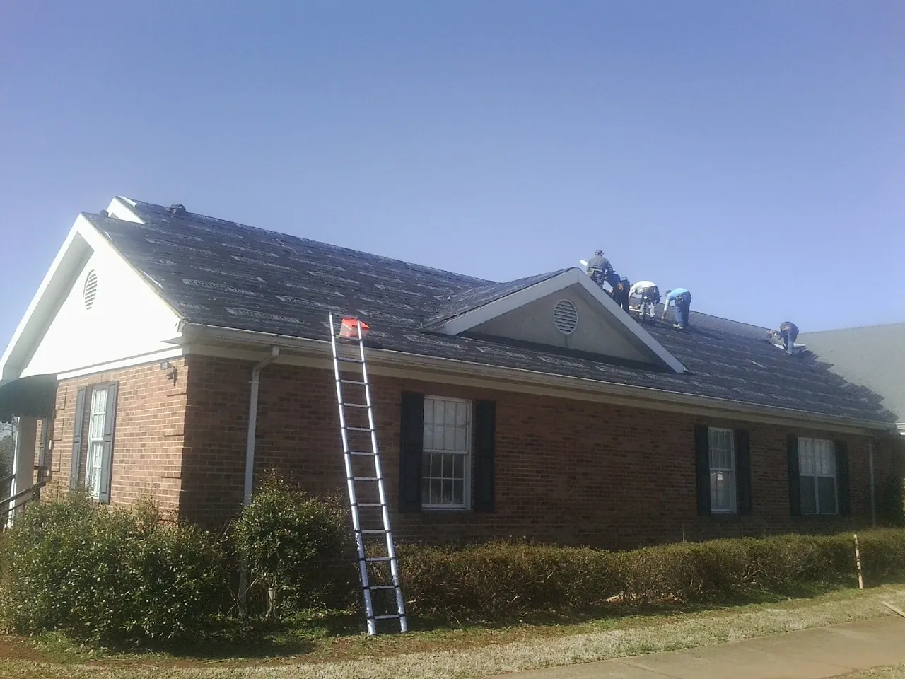 Crew tearing shingles off a brick ranch roof with a ladder set — Metal Roof Repair work by our crew in Fort Gratiot