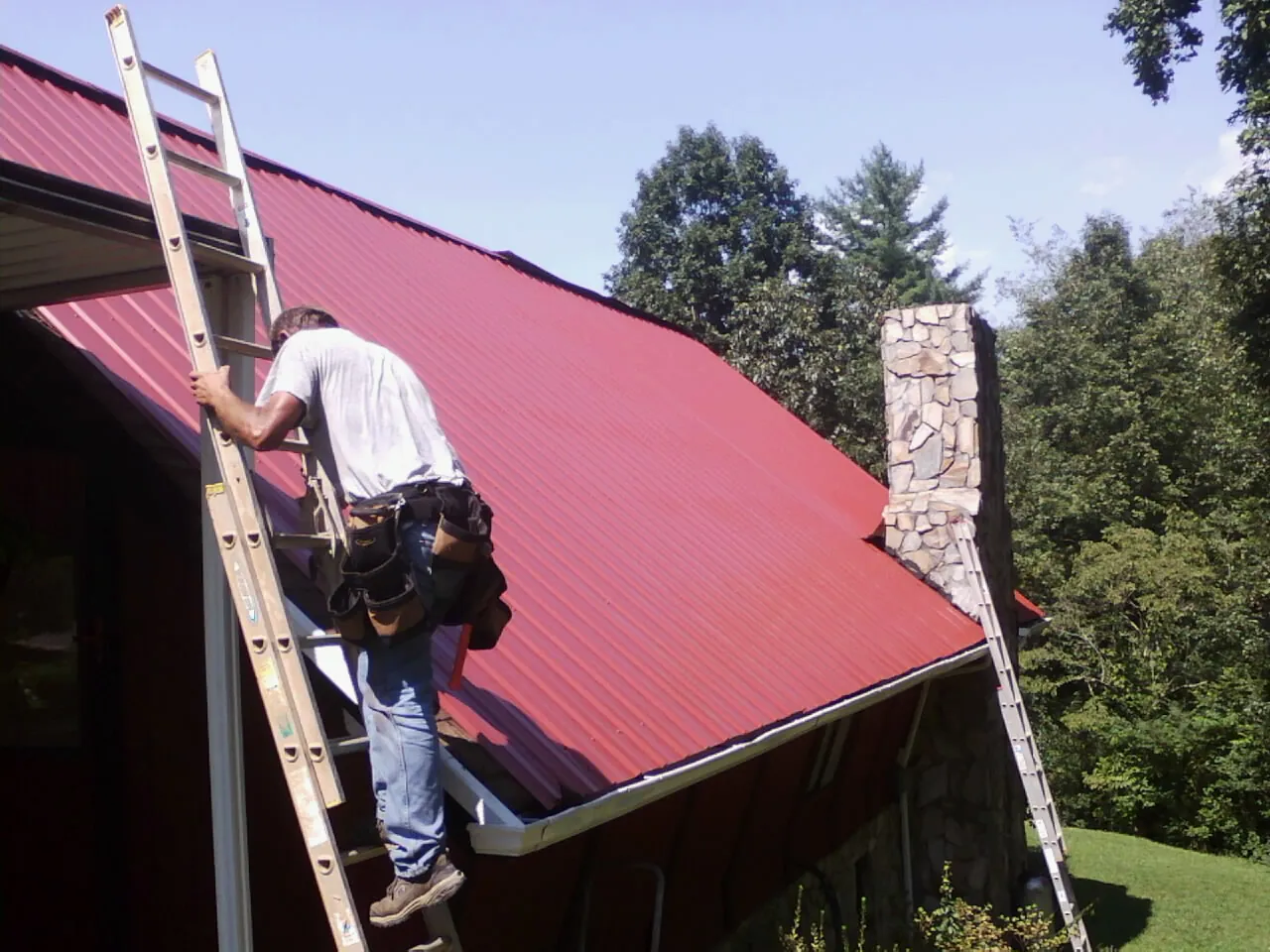 Roofer on a ladder beside a red metal roof and stone chimney — Soffit & Fascia Repair by Roofing Brothers in Fort Gratiot